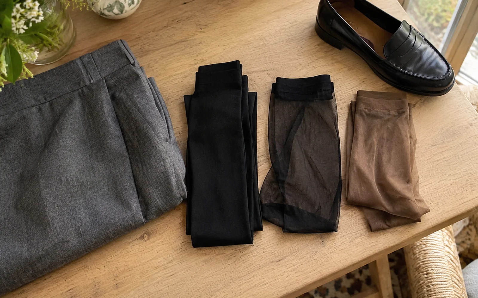 Trois paires de collants noirs opaques, noirs fins et bruns fins sur une table, près d’une jupe et d’un mocassin