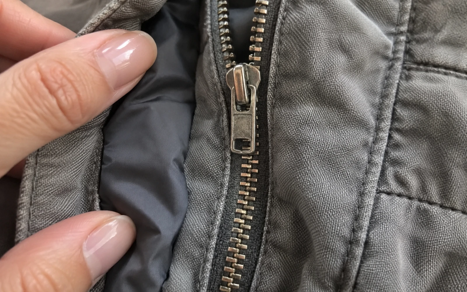 Deux doigts maintiennent le bord d’une veste grise près de sa fermeture.