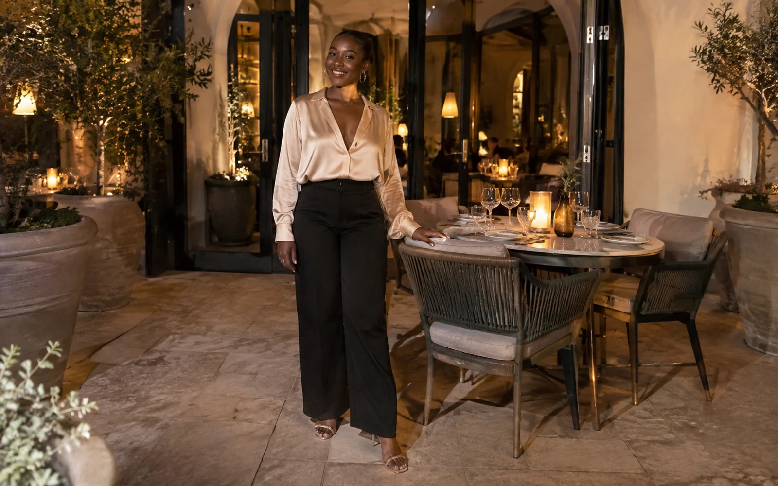Femme en pantalon noir fluide, blouse en satin champagne et sandales dorées près d’une table de restaurant